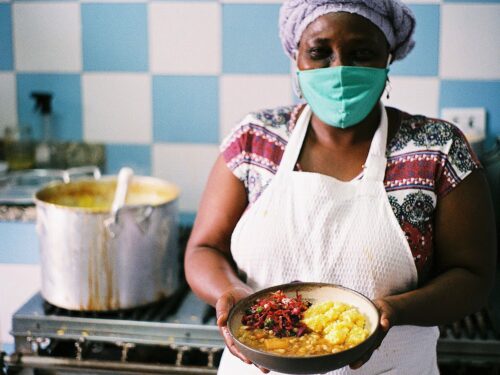Foto de uma mulher negra, em pé, em frente a um fogão, com um prato com comida nas mãos.