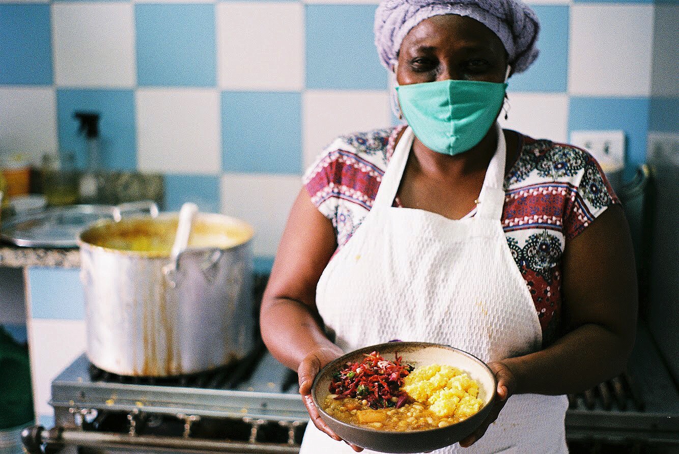 Foto de uma mulher negra, em pé, em frente a um fogão, com um prato com comida nas mãos.