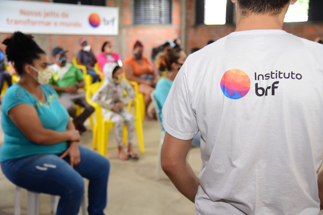 Foto de um homem, em pé, de costas e usando uma camiseta com o logo do Instituto BRF. À sua frente, há um grupo de pessoas sentadas.
